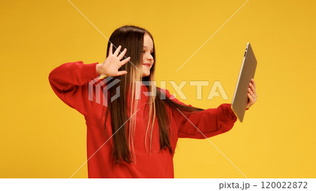 Girl in red sweater waving hello at tablet, during meeting online, smiling as she poses against yellow studio background. 120022872