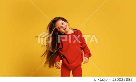 Joyful little girl in red sweater with loose hair, tilting her head and smiling against yellow studio background. 120022882