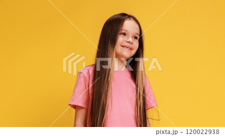 Pretty, charming girl, with long hair, dressed stylish attire looking at distance with smile against yellow studio background. 120022938