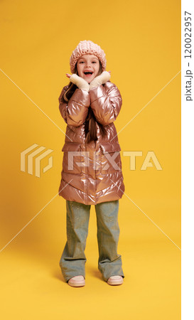 Happy little child, girl dressed winter clothes, smiling and holding hand near face against yellow studio background. Happy little child, girl dressed winter clothes, smiling and holding hand near face against yellow studio background. 120022957