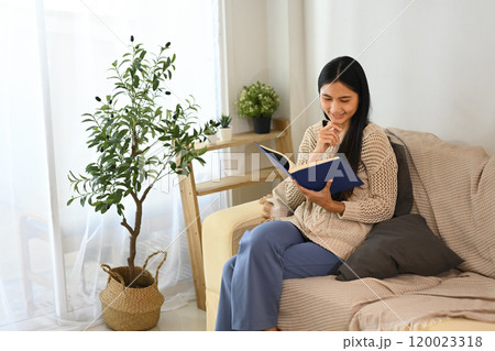 Beautiful young woman enjoys quiet time with a book in living room 120023318