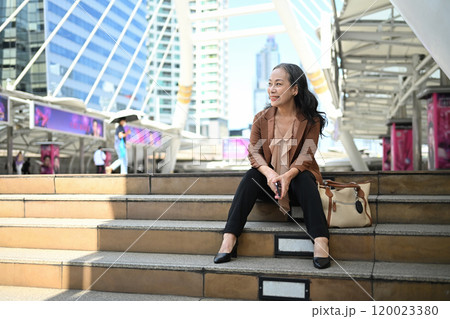 Confident mature businesswoman sitting on steps with a smile in the city center 120023380