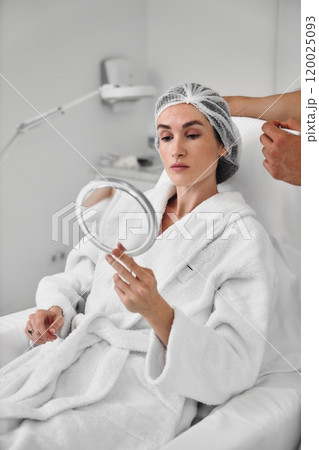 Woman in a spa robe examining her reflection in a mirror during treatment Woman in a spa robe examining her reflection in a mirror during treatment 120025093