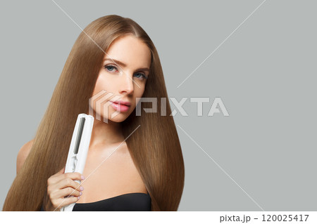 Brunette young female model woman with long brown smooth hairstyle holding straight iron.  120025417