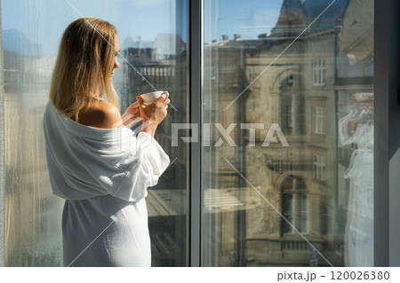Woman in elegant robe drinking coffee in hotel room and standing near window Woman in elegant robe drinking coffee in hotel room and standing near window 120026380