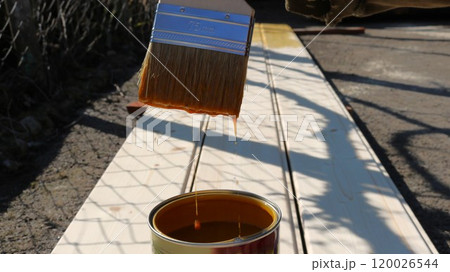 A 75-millimeter brush with orange paint or varnish dripping and dripping into a jar on the background of light wooden boards lying on asphalt 120026544