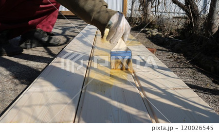 An employee wearing work clothes and gloves uses a brush to apply yellow paint or varnish to light-colored wooden boards that lie on the pavement. 120026545