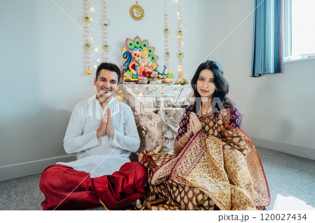 Smiling Indian couple dressed in traditional wear sitting at home altar, adorned with decors and offerings for god Ganesh after festive celebration. Indian culture, hindu ritual and customs. 120027434