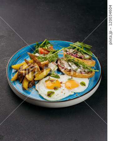 Breakfast plate with fried eggs, roasted potatoes, fresh salad, and sardine toasts on a blue dish Breakfast plate with fried eggs, roasted potatoes, fresh salad, and sardine toasts on a blue dish 120034604