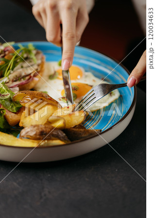 Close-up of hands cutting into a breakfast plate with roasted potatoes, eggs, and greens 120034633