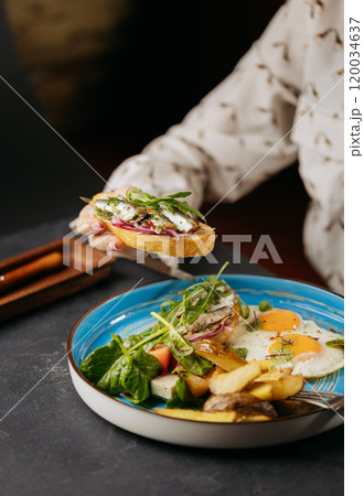 Hand holding sardine toast over a vibrant breakfast plate with eggs, salad, and potatoes Hand holding sardine toast over a vibrant breakfast plate with eggs, salad, and potatoes 120034637