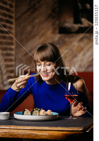 Smiling woman enjoying sushi with chopsticks, paired with a glass of rose wine in a cozy restaurant 120035117