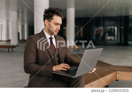 Serious focused businessman in stylish sharp brown suit working typing on laptop computer sitting outside by modern office building, showing determination and ambition in professional urban setting. 120035203