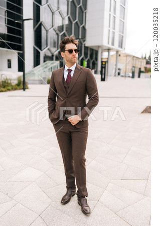 Full length vertical portrait of businessman in stylish brown suit standing in front of modern office building. Handsome serious business man in sunglasses posing outside holding hand in pocket. Full length vertical portrait of businessman in stylish brown suit standing in front of modern office building. Handsome serious business man in sunglasses posing outside holding hand in pocket. 120035218