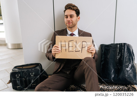 Portrait of poor handsome millennial businessman in suit sitting on city street holding sign HELP, illustrating hardships of poverty and unemployment. Concept of support in financial crises. 120035226