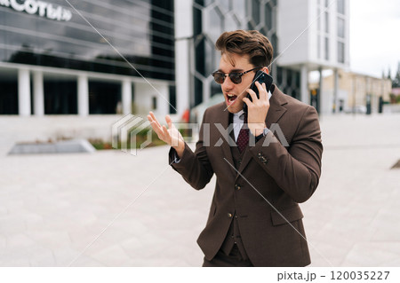 Portrait of mad screaming businessman in sunglasses and suit yelling on smartphone outside standing by office building, gesturing angrily with one hand. Concept of business communication. Portrait of mad screaming businessman in sunglasses and suit yelling on smartphone outside standing by office building, gesturing angrily with one hand. Concept of business communication. 120035227