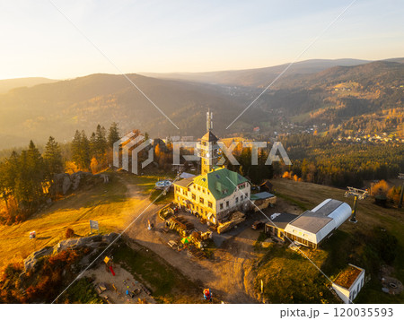The Tanvaldsky Spicak lookout tower stands prominently in the Jizera Mountains, surrounded by vibrant autumn foliage during a serene evening glow. Visitors are seen enjoying the view. 120035593