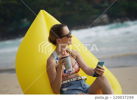 A young woman on the beach lounging on a yellow bean bag  120037289