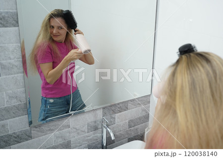 A woman is expertly styling her hair using a blow dryer and brush while in the bathroom A woman is expertly styling her hair using a blow dryer and brush while in the bathroom 120038140