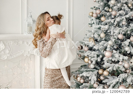 Blonde mother and little daughter in studio posing for a photo. Woman wearing golden dress and daughter white one. Family celebrating Christmas together. 120042789