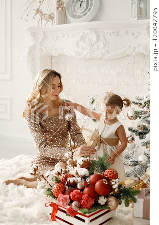 Blonde mother and little daughter in studio posing for a photo. Woman wearing golden dress and daughter white one. Family decorating Christmas together. 120042795