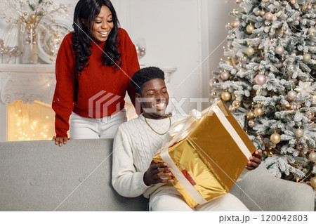 Young black couple with gifts in Christmas eve. Man sitting on a coach at home. Brunette woman standing behind and presented a gift for her boyfriend. 120042803