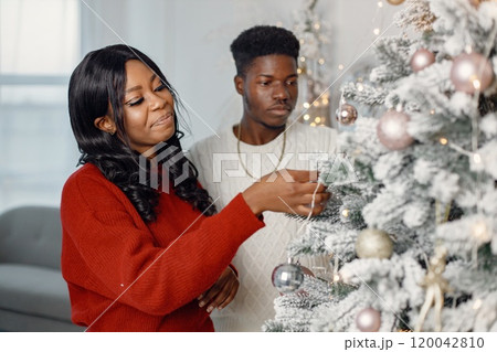 Happy black couple decorating Xmas tree together. Man amd woman celebrating winter holidays at home. Brunette woman wearing a red sweater. Happy black couple decorating Xmas tree together. Man amd woman celebrating winter holidays at home. Brunette woman wearing a red sweater. 120042810