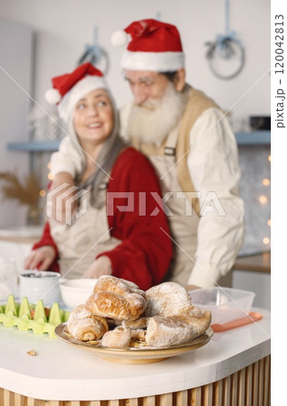Senior man and his wife baking croissants together. Old bearded man standing together near his wife with grey hair. Man wearing white sweater and woman red one. 120042813