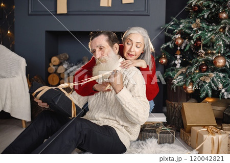 Senior man celebrating christmas with his wife near beautiful christmas tree. Old bearded man sitting on a floor near his wife with grey hair and holding a gifts. Man wearing white sweater and woman Senior man celebrating christmas with his wife near beautiful christmas tree. Old bearded man sitting on a floor near his wife with grey hair and holding a gifts. Man wearing white sweater and woman 120042821