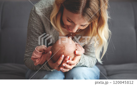 Close up happy mother tenderly holding and embracing small baby, gazing lovingly while sitting on dark grey sofa at home. Mom with newborn indoors 120045812