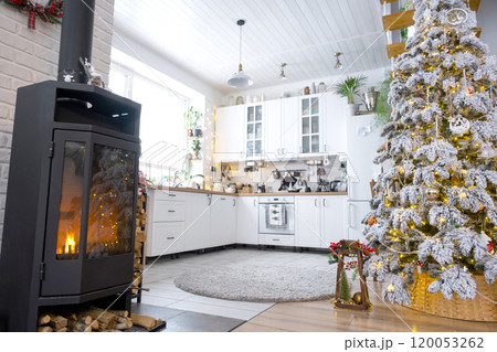 Festive white modern interior of house is decorated for Christmas and New Year in loft style with black stove, fireplace, Christmas tree. Potted plants, firewood in the woodpile 120053262