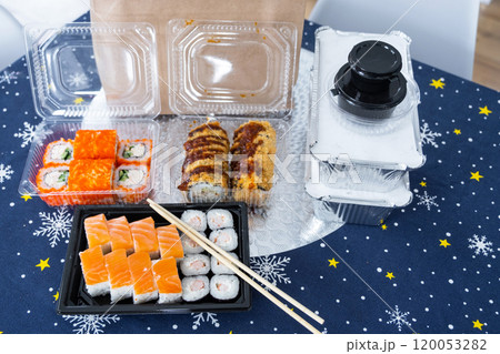 Delivery of sushi and rolls set top view on the table with a Christmas decor with a Christmas tree and a tablecloth with snowflakes, home cooking 120053282
