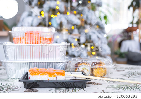 Delivery of sushi and rolls in food containers on the table in the festive interior of the kitchen of the house with a white Christmas tree for Christmas and New year. Food delivery for the holidays 120053293