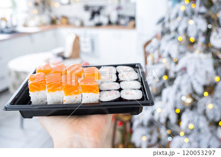 Delivery of sushi and rolls set is hold in hand in the festive interior of house with a white Christmas tree for Christmas and New year. Food delivery for the holidays 120053297