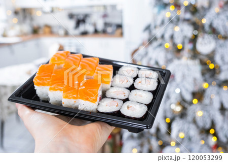 Delivery of sushi and rolls set is hold in hand in the festive interior of house with a white Christmas tree for Christmas and New year. Food delivery for the holidays 120053299