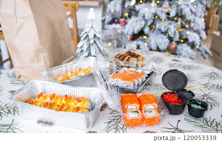 Delivery of sushi and rolls in food containers on the table in the festive interior of the kitchen of the house with a white Christmas tree for Christmas and New year. Food delivery for the holidays Delivery of sushi and rolls in food containers on the table in the festive interior of the kitchen of the house with a white Christmas tree for Christmas and New year. Food delivery for the holidays 120053339