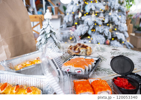 Delivery of sushi and rolls in food containers on the table in the festive interior of the kitchen of the house with a white Christmas tree for Christmas and New year. Food delivery for the holidays 120053340