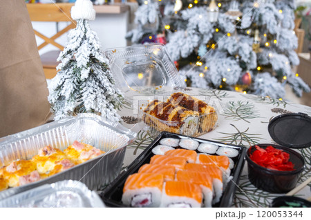 Delivery of sushi and rolls in food containers on the table in the festive interior of the kitchen of the house with a white Christmas tree for Christmas and New year. Food delivery for the holidays 120053344