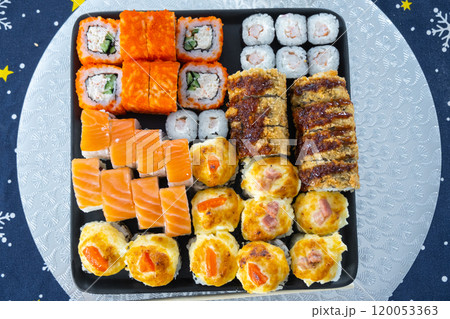 Delivery of sushi and rolls set top view on the table with a Christmas decor with a Christmas tree and a tablecloth with snowflakes, home cooking 120053363