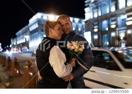 A couple walks hand-in-hand through a city at night, the man holding a bouquet of flowers A couple walks hand-in-hand through a city at night, the man holding a bouquet of flowers 120053500