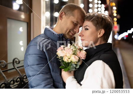 A couple walks through the city at night, holding a bouquet of pink flowers A couple walks through the city at night, holding a bouquet of pink flowers 120053553