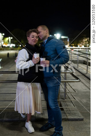 A couple strolls through the city at night, holding warm drinks as they enjoy each other's company 120053580