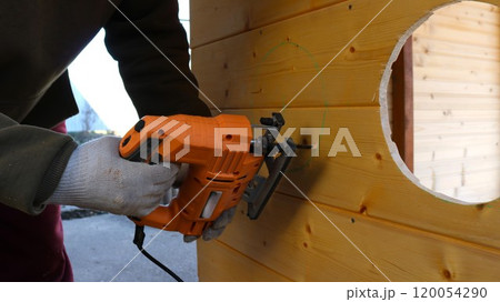 A man in work clothes and gloves uses a jigsaw to cut a wooden wall according to markings. Making round holes in a wooden partition with an orange jigsaw A man in work clothes and gloves uses a jigsaw to cut a wooden wall according to markings. Making round holes in a wooden partition with an orange jigsaw 120054290