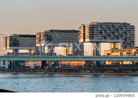 A modern cityscape featuring three unique crane-shaped buildings along the river, with a bridge in front carrying a train. The buildings reflect the warm light of the setting sun. 120054868