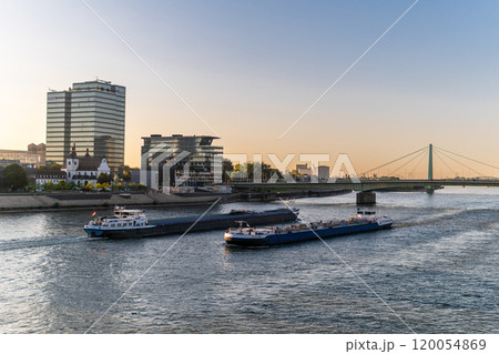 Cargo ships, barges transport goods on the Rhine in Cologne. Navigable river. Modern glass houses on the river bank. Beautiful sunset. Metal bridge over the river. Beautiful church. Blue sky. 120054869