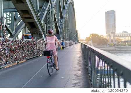 A woman on a bicycle rides on a bridge over the Rhine in the city of Cologne. A beautiful metal bridge at dawn. Many locks are attached to the bridge. The woman in a red dress and a red helmet. A woman on a bicycle rides on a bridge over the Rhine in the city of Cologne. A beautiful metal bridge at dawn. Many locks are attached to the bridge. The woman in a red dress and a red helmet. 120054870