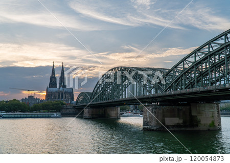 View of Cologne Cathedral and Hohenzollern Bridge with a cargo ship and passenger boat navigating the Rhine River under a clear blue sky, showcasing Cologne's iconic cityscape and vibrant river life. 120054873