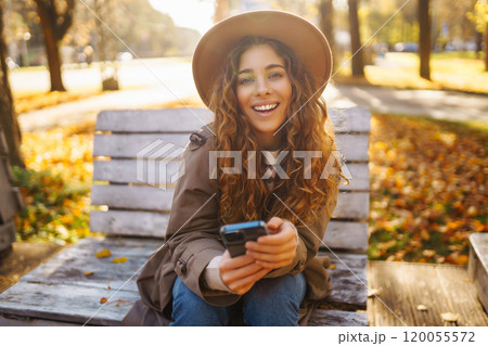 Young smiling woman sitting on bench in autumn park using phone in stylish clothes happy mood. Young smiling woman sitting on bench in autumn park using phone in stylish clothes happy mood. 120055572