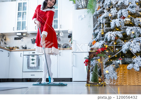 A woman in a red dress and Santa hat cleans the floor with a mop in a white kitchen with Christmas decor and a Christmas tree. Cleaning the house and preparing for the holidays Christmas and New Year A woman in a red dress and Santa hat cleans the floor with a mop in a white kitchen with Christmas decor and a Christmas tree. Cleaning the house and preparing for the holidays Christmas and New Year 120056432