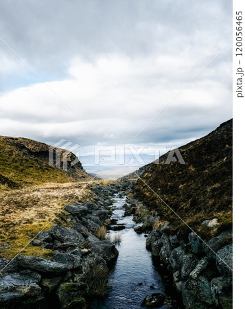 A small river cuts through a valley, flanked by rocky slopes. The sky is overcast, and the distant landscape is partially visible through the cloud cover. 120056465